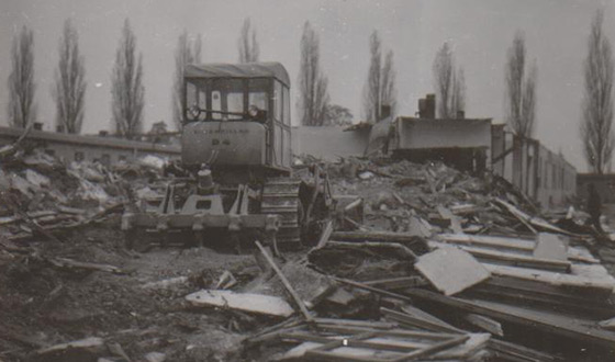 A bulldozer crosses the remains of a demolished barrack and heads towards a part of the barrack still standing. (Dachau Concentration Camp Memorial Site)