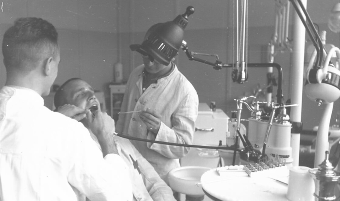 La photographie montre un détenu soigné par deux dentistes dans une salle de soins dentaires dotée d’un équipement moderne pour l’époque.