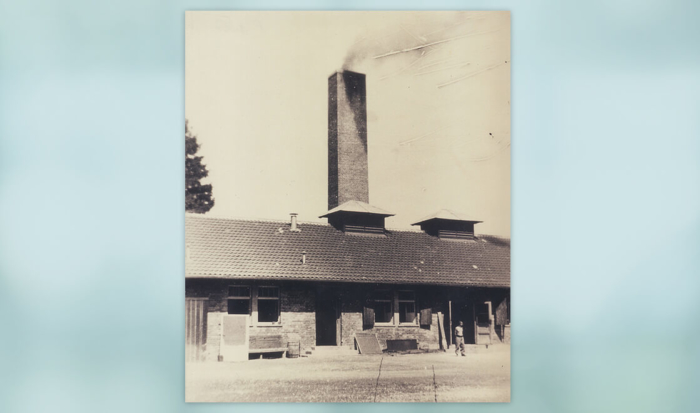 The photo shows the part of “barrack X” with the large chimney, dark smoke billowing out into the sky. A man dressed only in pants and shoes is standing in front of the building; all of the doors and windows are wide open.