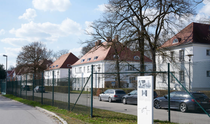 Hinter einem Maschendrahtzaun ist eine Reihe von mehrstöckigen Häusern zu erkennen, die im Villen-Stil erbaut wurden. Vor dem Zaun ist eine Informationstafel angebracht, die die Geschichte dieser Bauten aufgreift.