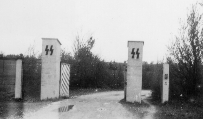 La photographie montre deux hauts poteaux carrés en pierre sur lesquels a été apposé l’emblème des SS. Entre ces poteaux, le portail est ouvert.