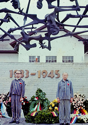 Two former prisoners in striped prisoners’ garb stand in front of the International Monument; several wreaths are behind them, leaning against the wall on which the dates “1933-1945” are mounted. (Daniel Gordana and Gabriel Glid)