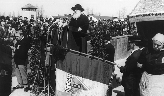 In front of the Jewish memorial Rabbi David Spiro stands on a podium and recites the prayer for the dead into several microphones. A large crowd is gathered in the background.