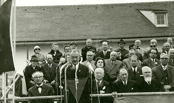 View of a stand in front of the former maintenance building. Around 30 persons sit solemnly while a man stands at a lectern and speaks into a microphone. (CID)