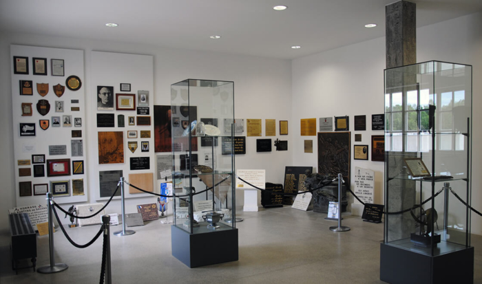 La photographie présente une vue de la salle du souvenir. Aux murs sont apposées diverses plaques commémoratives de couleurs et de formes différentes, en mémoire de victimes individuelles ou bien de groupes de victimes. Deux hautes vitrines en verre placées au milieu de la salle renferment d’autres plaques et objets commémoratifs.