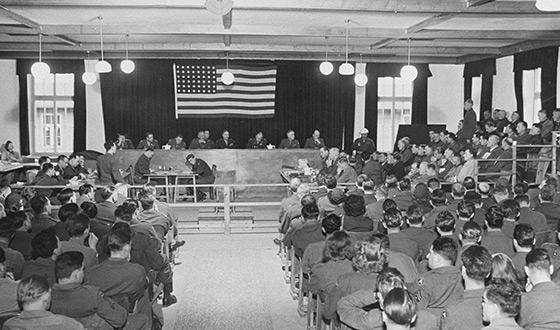 Court room seen from the rear: behind the bench at the front, eight judges sit beneath the U.S. flag. To their right on the photo are some 20 defendants, while court clerks sit in front of the judges’ bench. The spectator seats are full. (USHMM)