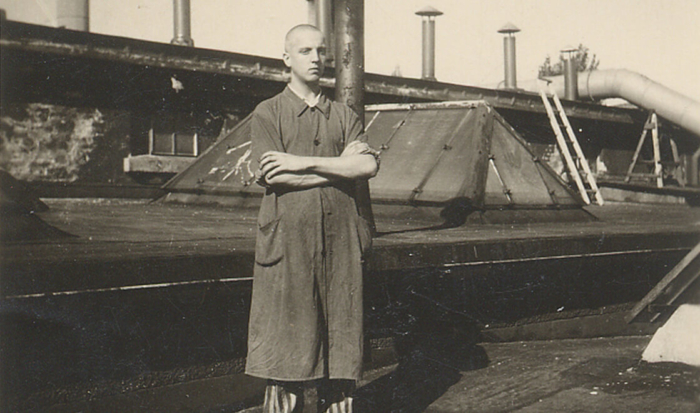 A prisoner stands with folded arms on the roof of a greenhouse. He is wearing the striped prisoner clothing underneath a work coat.