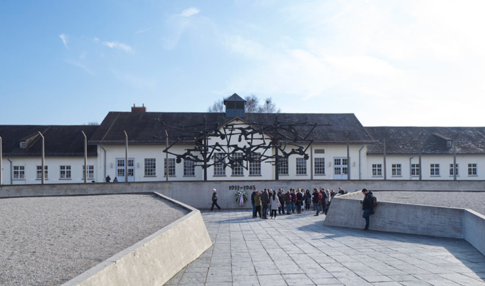 The photo shows the broad path leading to the International Monument. Mounted on a wall with the inscription 1933-1945 is a bronze sculpture of human figures entangled in barbed wire. Behind them are the stylized concrete pillars symbolizing the guard installation.