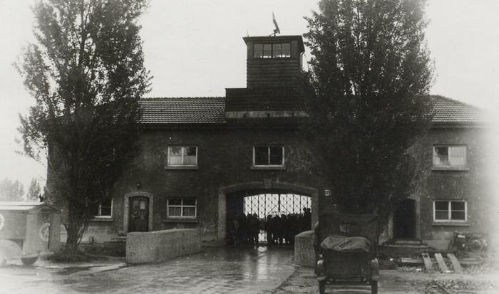 Derrière deux arbres, donnant sur le camp, se dresse le bâtiment tout en longueur du Jourhaus, au milieu duquel se trouve une large arcade. Un portail en fer forgé portant l’inscription « Arbeit macht frei » servait d’entrée et de sortie au camp et est, encore aujourd’hui, l’accès principal pour les visiteurs du mémorial.