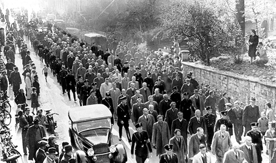 Scores of men in coats walk down a street in Baden-Baden, escorted by SS men in uniform – onlookers line the street, SS propaganda photo (Federal archives)