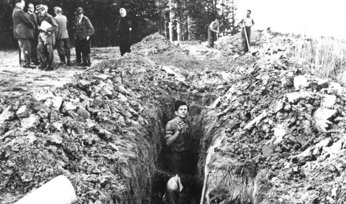 En la foto se ve un hombre que está de pie hasta los hombros en una fosa excavada a lo largo. A dcha. e izq. de la fosa hay tierra y huesos amontonados.