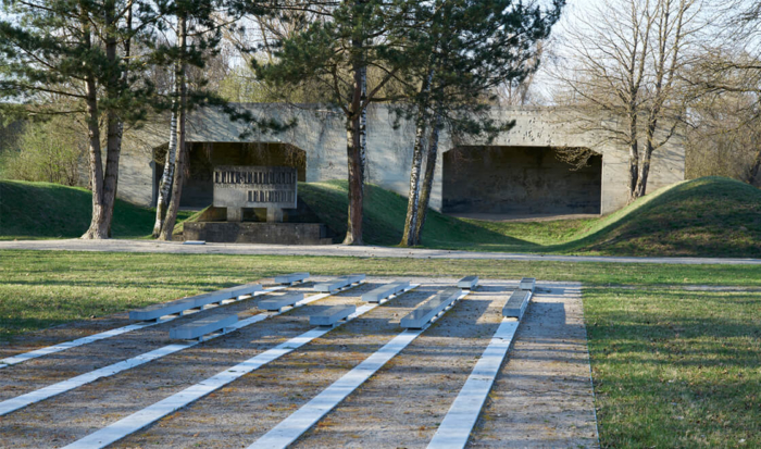 Hidden somewhat by trees, two shooting stands are located in the background. The shooting lanes are bordered on the left and right by the grassy mounds. In front of the shooting stands is a stone monument dedicated to the murdered Soviet prisoners of war. In the foreground are the long rows of parallel stone slabs, embedded into the ground. Mounted on some of the slabs are commemorative plaques for the identified victims amongst the Soviet prisoners of war.