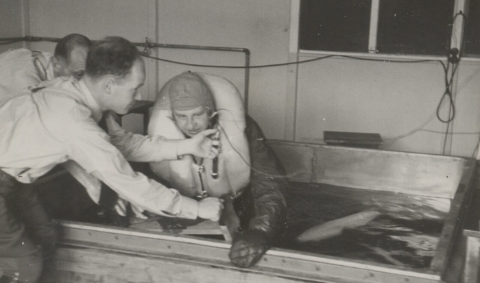 Wrapped up in thick clothes and wearing a life vest, a prisoner sits in a shallow water basin. While forced to endure the life-threatening hypothermia experiment, two SS men stand next to the basin. Leaning forward they are measuring the prisoner’s responses to the extreme cold.