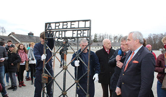 Press briefing on the former roll call area. The postern with the historical inscription “Arbeit macht frei” is placed upright for photographers. The gate is meanwhile corroded. Standing beside the gate are Jean Michel Thomas, the president of the CID, Karl Freller, the Foundation director, and Dr Ludwig Spaenle, Culture Minister.