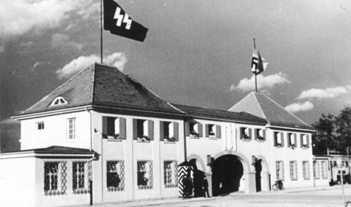 A striking building with a gateway, a guard stationed at the entrance and flags with SS and swastika symbols.