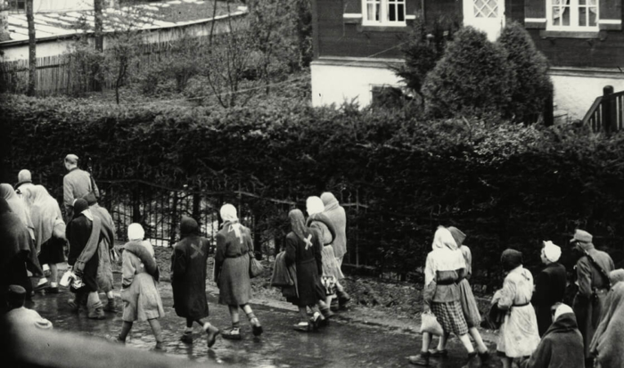 Women prisoners on a march are photographed from a window. They are wearing scarfs to protect themselves from the rain. Many of them also have wrapped blankets around their bodies, which they will then use for camping at night. An armed SS man is to be seen on the left.