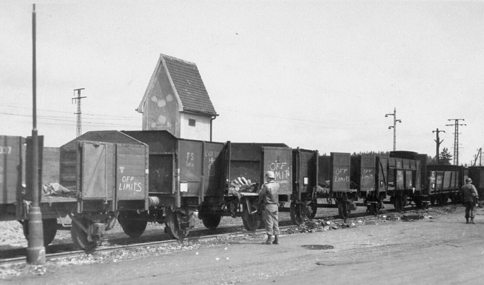 En la foto se ve un tren parado con vagones abiertos. Todas las puertas del tren están abiertas y en algunos vagones hay cadáveres. Un soldado estadounidense está de pie delante del tren y observa el terrible hallazgo.