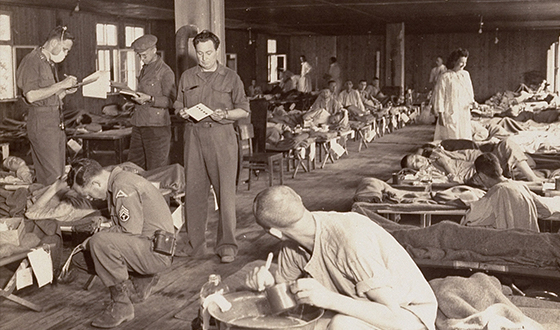 Więźniowie w szpitalnych koszulach na pryczach. Opiekują się nimi amerykańscy żołnierze (USHMM)