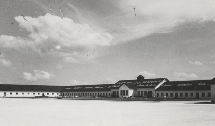 The black-and-white photograph shows the maintenance building. In 1939 a quote from Heinrich Himmler was painted in large white letters on the side of the maintenance building roof facing the roll call area and the barracks: “There is one path to freedom. Its milestones are: obedience, honesty, cleanliness, sobriety, diligence, order, self-sacrifice, truthfulness, love of the fatherland.”