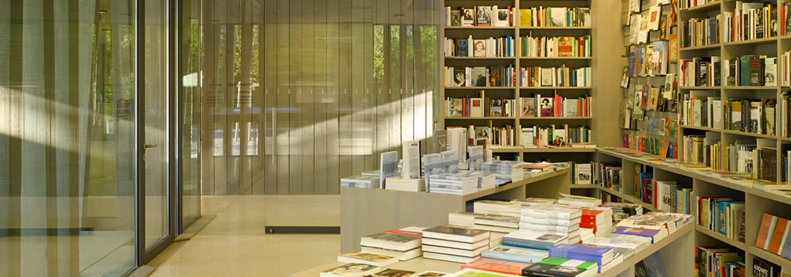 Foto della libreria “Literaturhandlung” in cui si vedono libri esposti sui tavoli e negli scaffali; la libreria si trova all'interno del centro visitatori del memoriale del campo di concentramento di Dachau