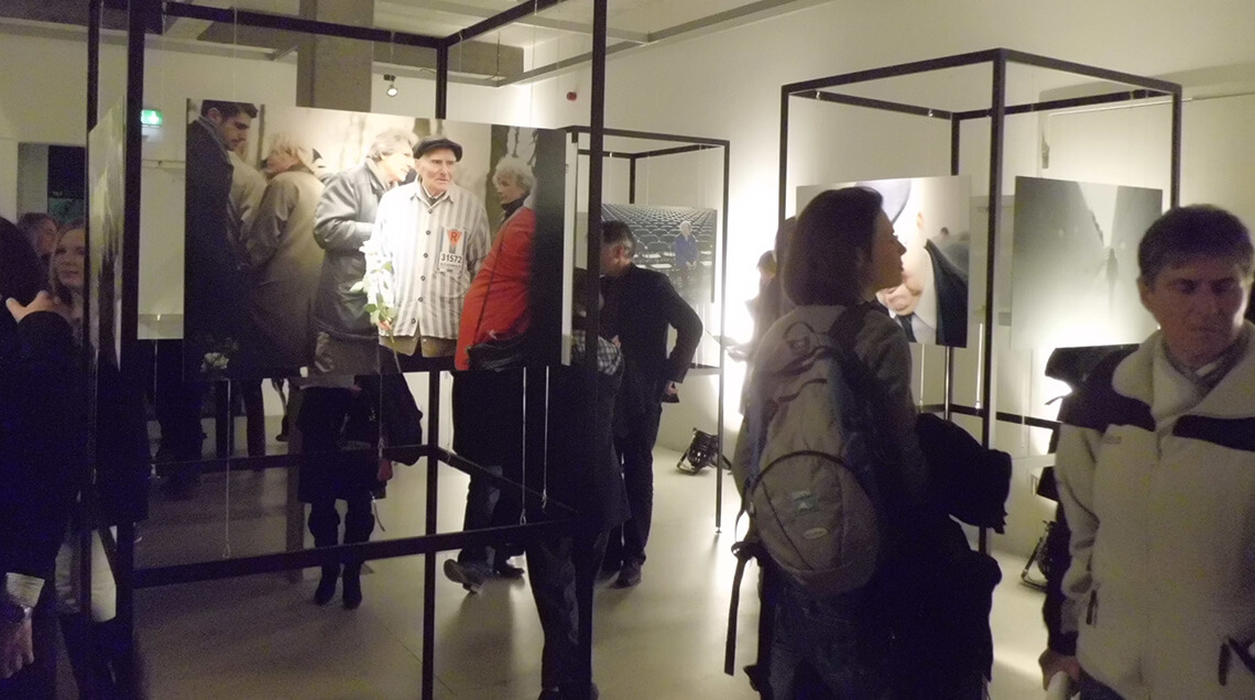 Besucher in der Doppelausstellung „Generationen” und „Blickwinkel” betrachten Fotografien. Mehrere würfelförmige Metallhalterungen stehen im Raum verteilt, an diesen sind Bilder befestigt, die ehemalige Häftlinge zeigen, die an die Orte ihres Leidens zurückkehren.