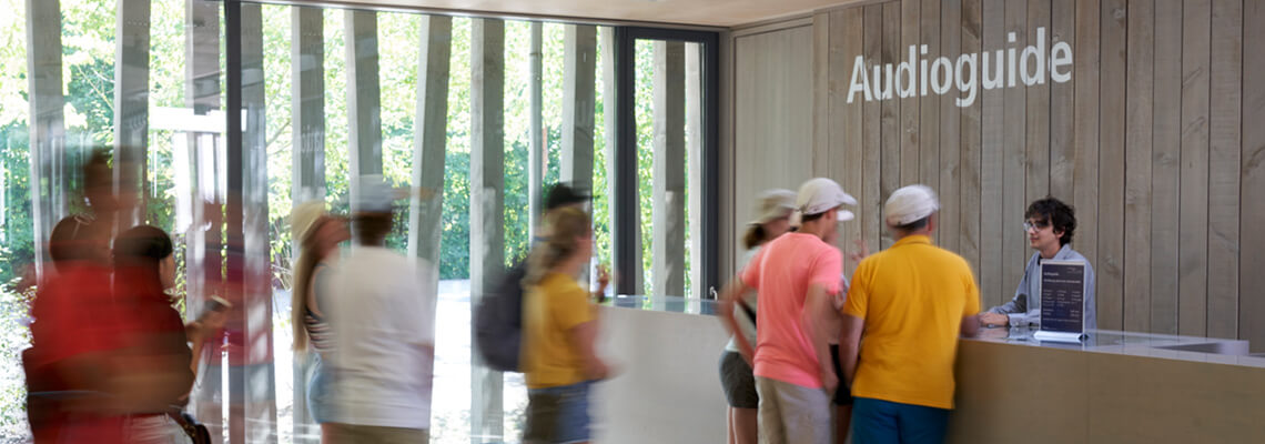 Besucherinnen und Besucher vor der Theke des Audioguide-Verleihs im Besucherzentrum der KZ-Gedenkstätte Dachau