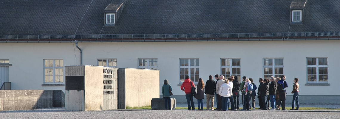 Grupa zwiedzających stoi przy Pomniku Narodów przed dawnym budynkiem gospodarczym