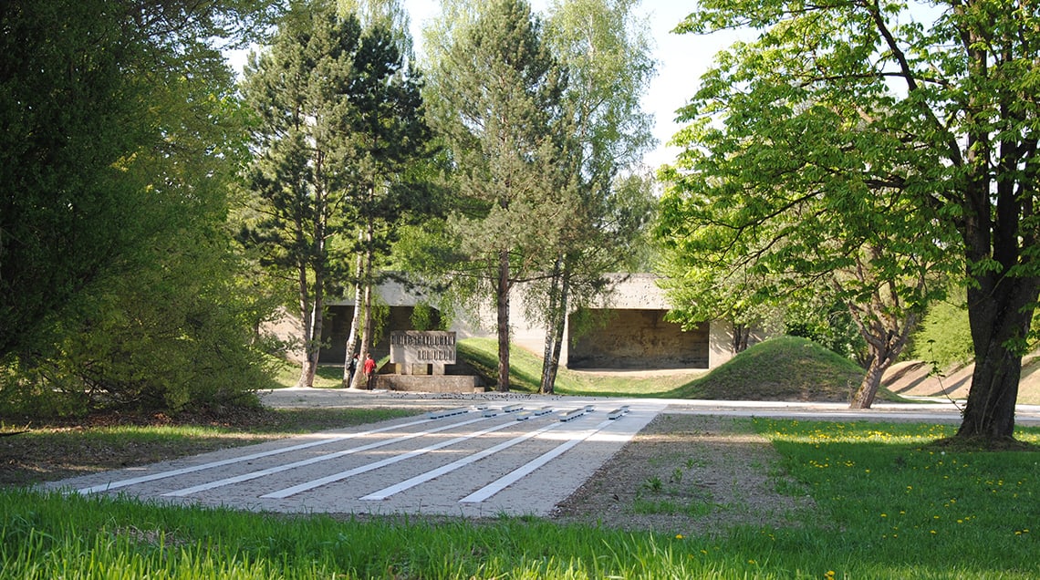 Zwei Besucher stehen vor dem Mahnmal auf dem ehemaligen “SS-Schießplatz Hebertshausen”, rechts von ihnen ist der Kugelfang aus Beton zu sehen.