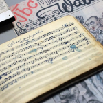 Abgebildet sind Werke, die heimlich im KZ Dachau entstanden sind: ein mit Kompositionen gefülltes Notenheft und eine gezeichnete Einladung zu einem Konzert