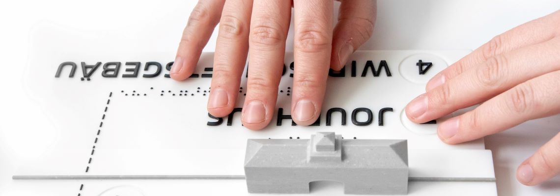 Finger ertasten ein taktiles Modell mit dem Jourhaus und Brailleschrift.