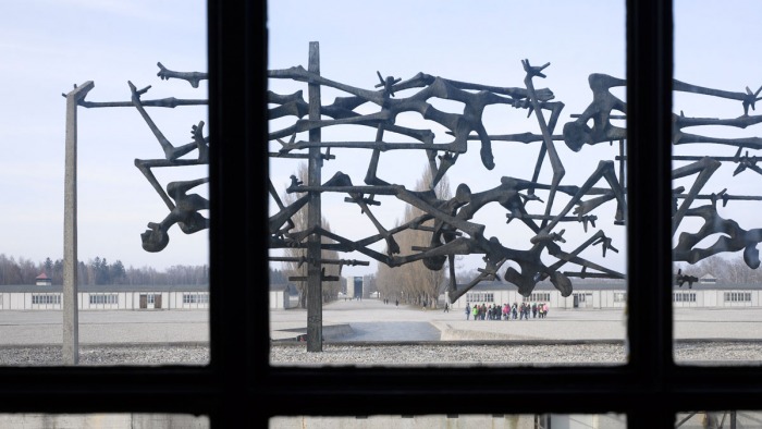 Durch ein Fenster fotografiert, ist auf dem Bild das Interantionale Mahmal zu sehen, im Hintergrund zwei rekonstruierte Baracke und die ehemalige Lagerstraße.