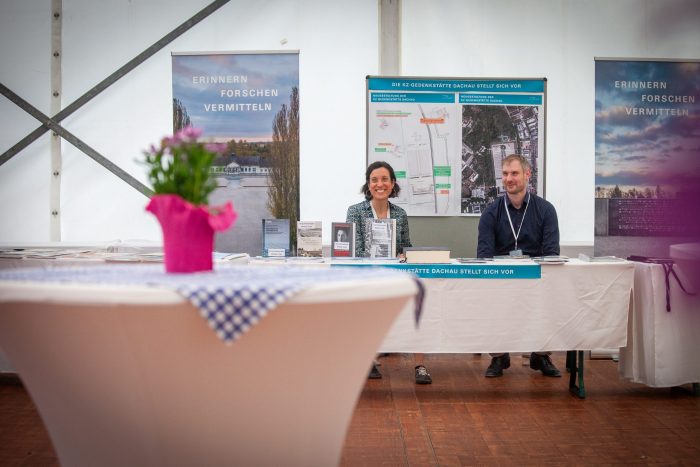 Zwei Personen sitzen an einem Tisch und lächeln, im Hintergrund ist Infomaterial der KZ-Gedenkstätte Dachau zu sehen
