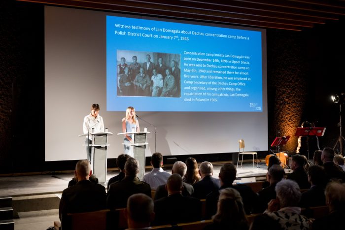 Zwei Personen stehen vor einer großen Leinwand auf einer Bühne und lesen Text vor