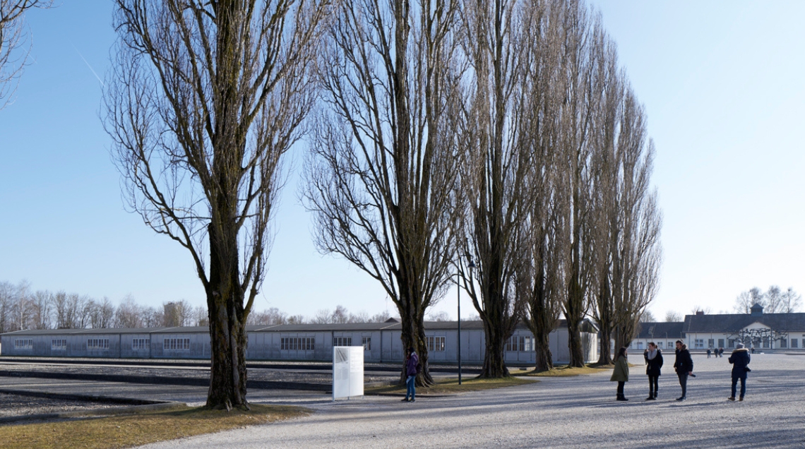 Blick auf das Gelände der Gedenkstätte mit rekonstruierter Baracke, ehemaliger Lagerstraße mit Besuchenden und ehemaligem Wirtschaftsgebäude