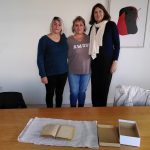 Drei Frauen stehen vor einem Tisch, auf dem ein erkennbar altes Buch auf einem Tuch ausgebreitet liegt